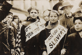 Two young women wearing protest sashes look at the camera. They hold small American flags with men and boys standing around them.