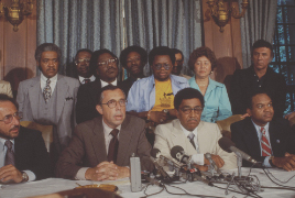 Two Jewish men and two Black men in suits sit at a table with microphones in front of them. Several men and one woman stand in the background.