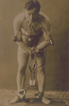 Harry Houdini stands looking at the camera, wearing a loincloth and wrapped in chains with heavy locks.