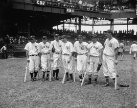 Seven baseball players in their uniforms (New York Yankees, Detroit Tigers, and Boston Red Sox) stand in a row leaning on their bats with people gathering in the stands behind them.